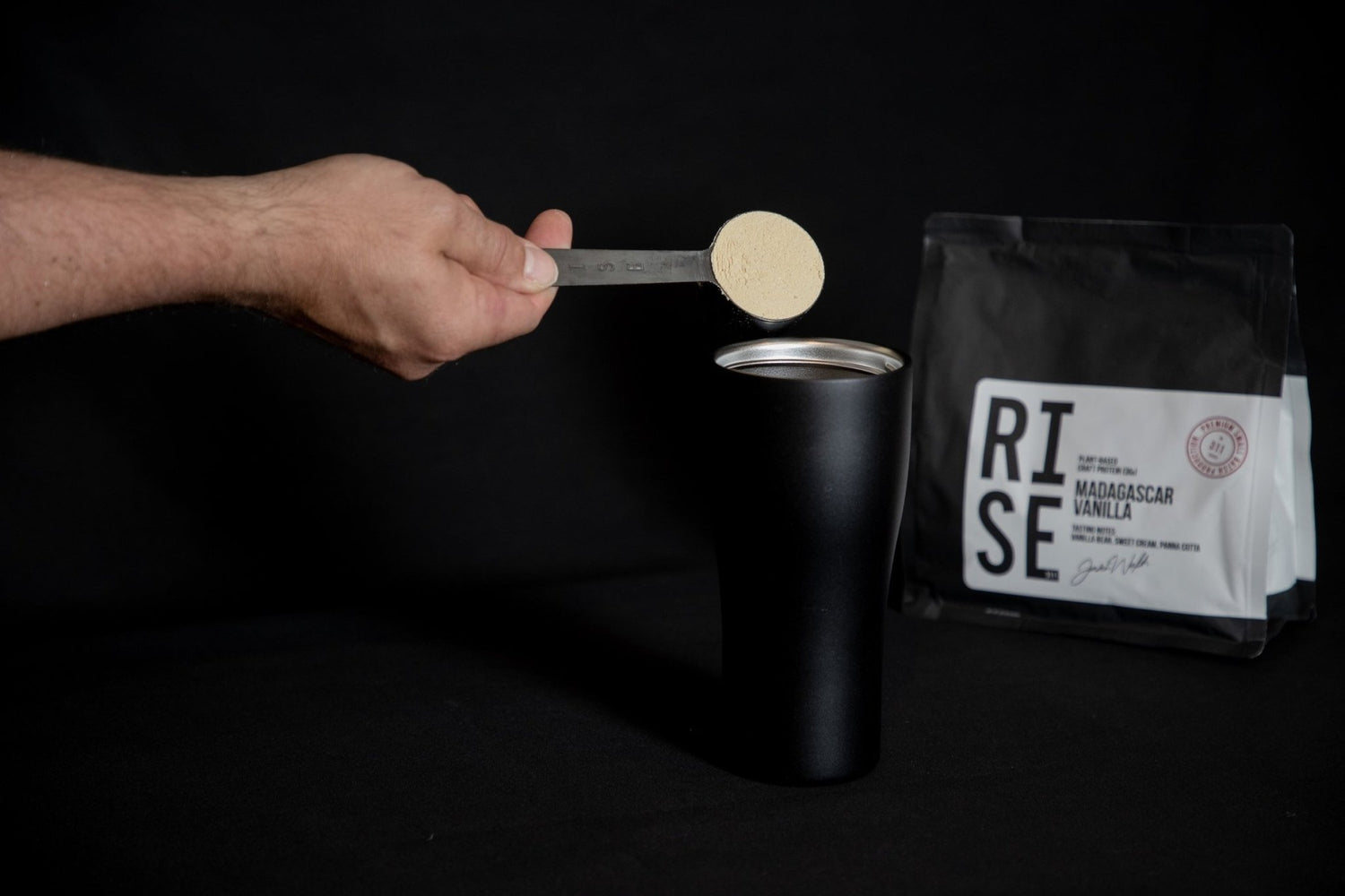 Hand holding stainless steel protein scoop filled with RISE vanilla protein powder.
