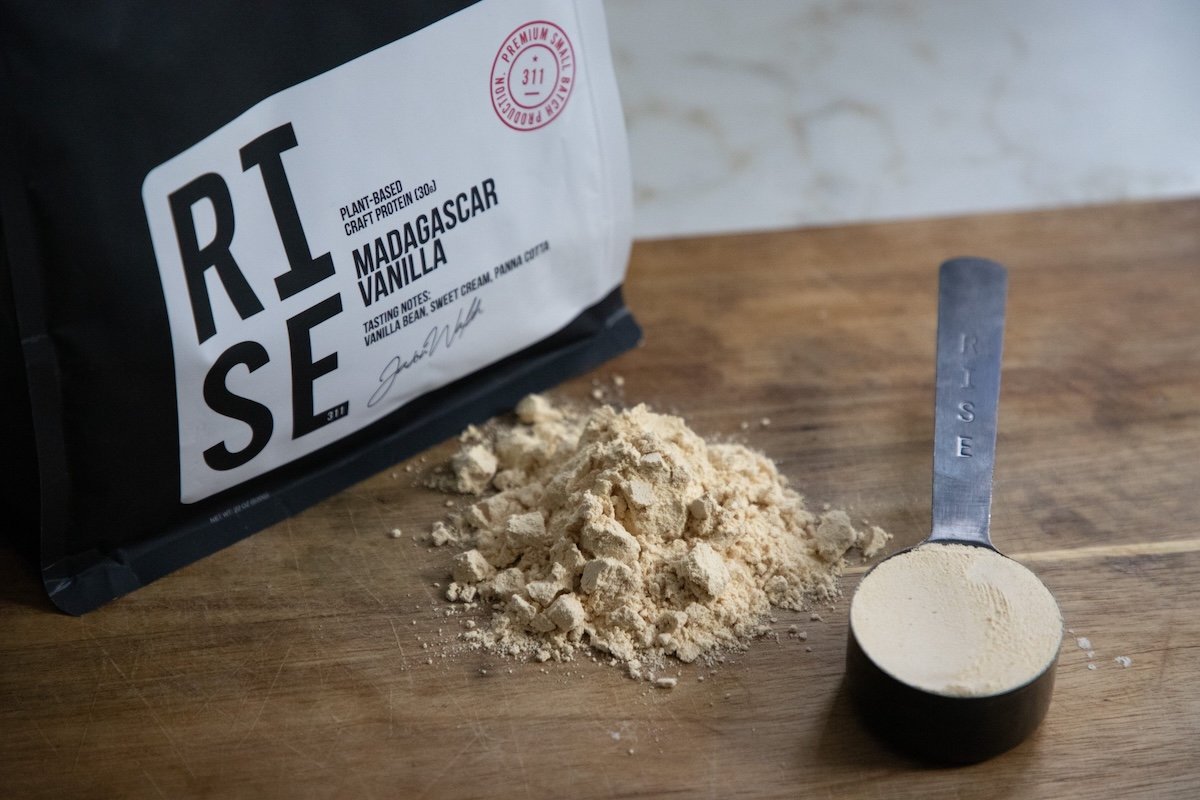 RISE vanilla protein pouch with stainless scoop on a wooden bench (overhead shot)
