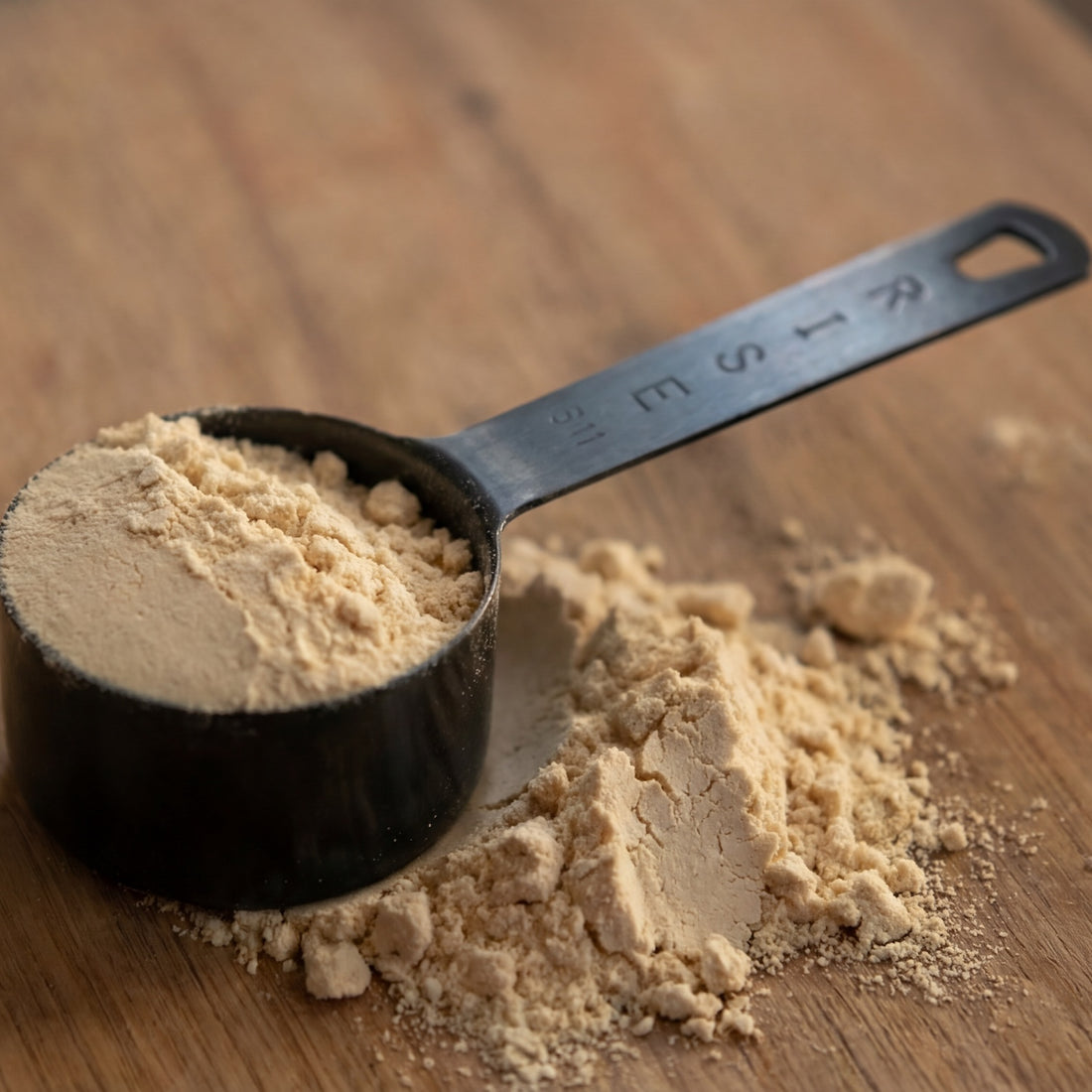 Stainless steel protein scoop shown filled with powder and resting on a wooden surface.