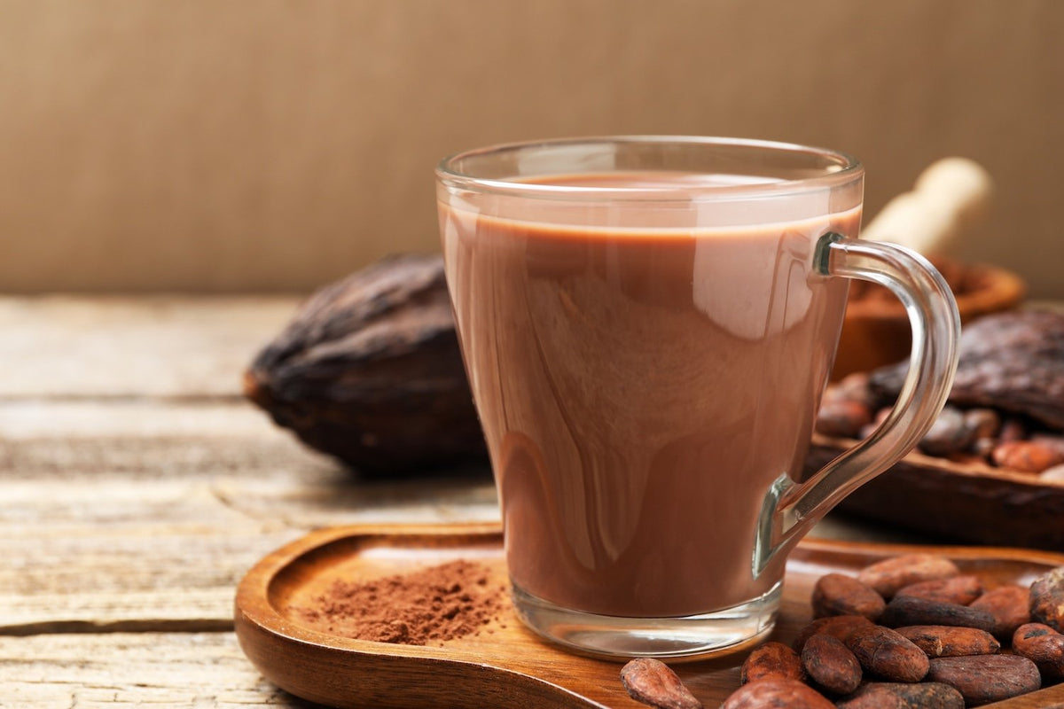 A clear glass mug filled with chocolate protein powder sitting on a wooden tray, surrounded by cocoa beans and cocoa powder.